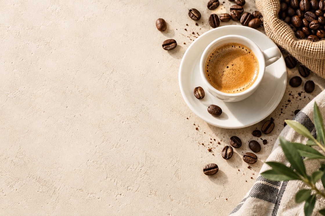 Espresso in einer weißen Tasse auf einem hellen Steintisch, daneben verstreute Kaffeebohnen und ein Jutesack